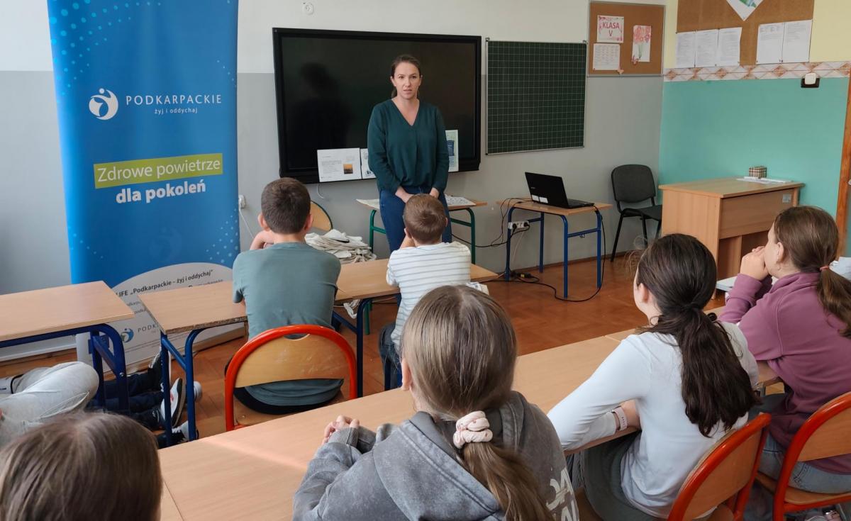 Nauczycielka prowadzi lekcję dla uczniów w klasie. Na ścianie wisi plakat "Zdrowe powietrze dla pokoleń".